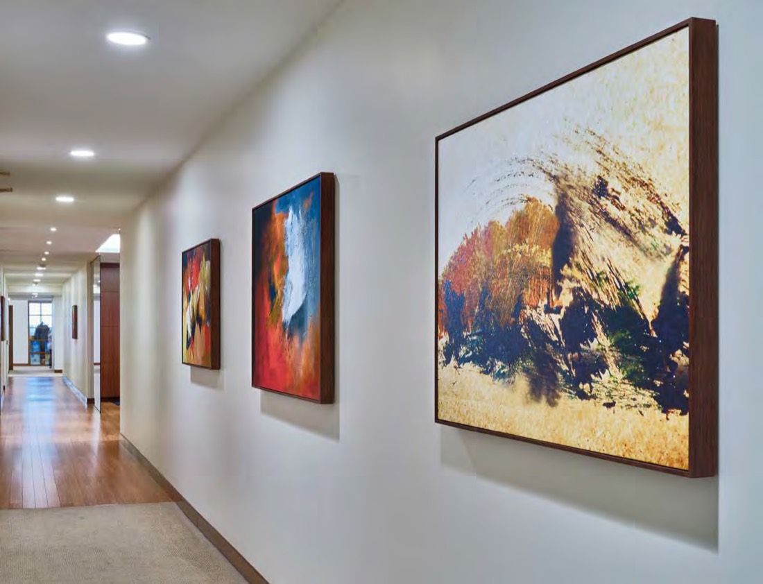 Framed paintings of increasing size hung along a white office corridor with wood flooring